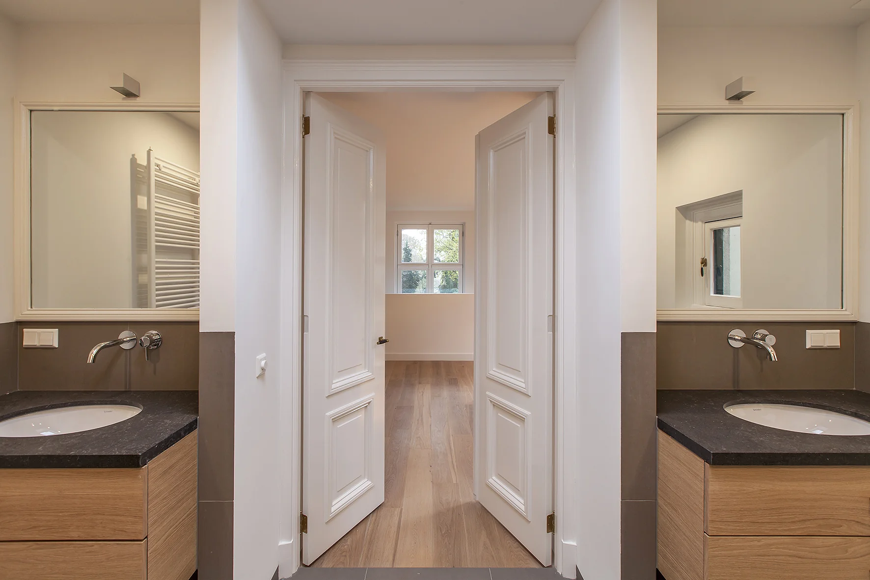 Moderne badkamer met dubbele wastafel, houten ladekasten en openslaande deuren naar een lichte kamer aan de Naarderstraat.