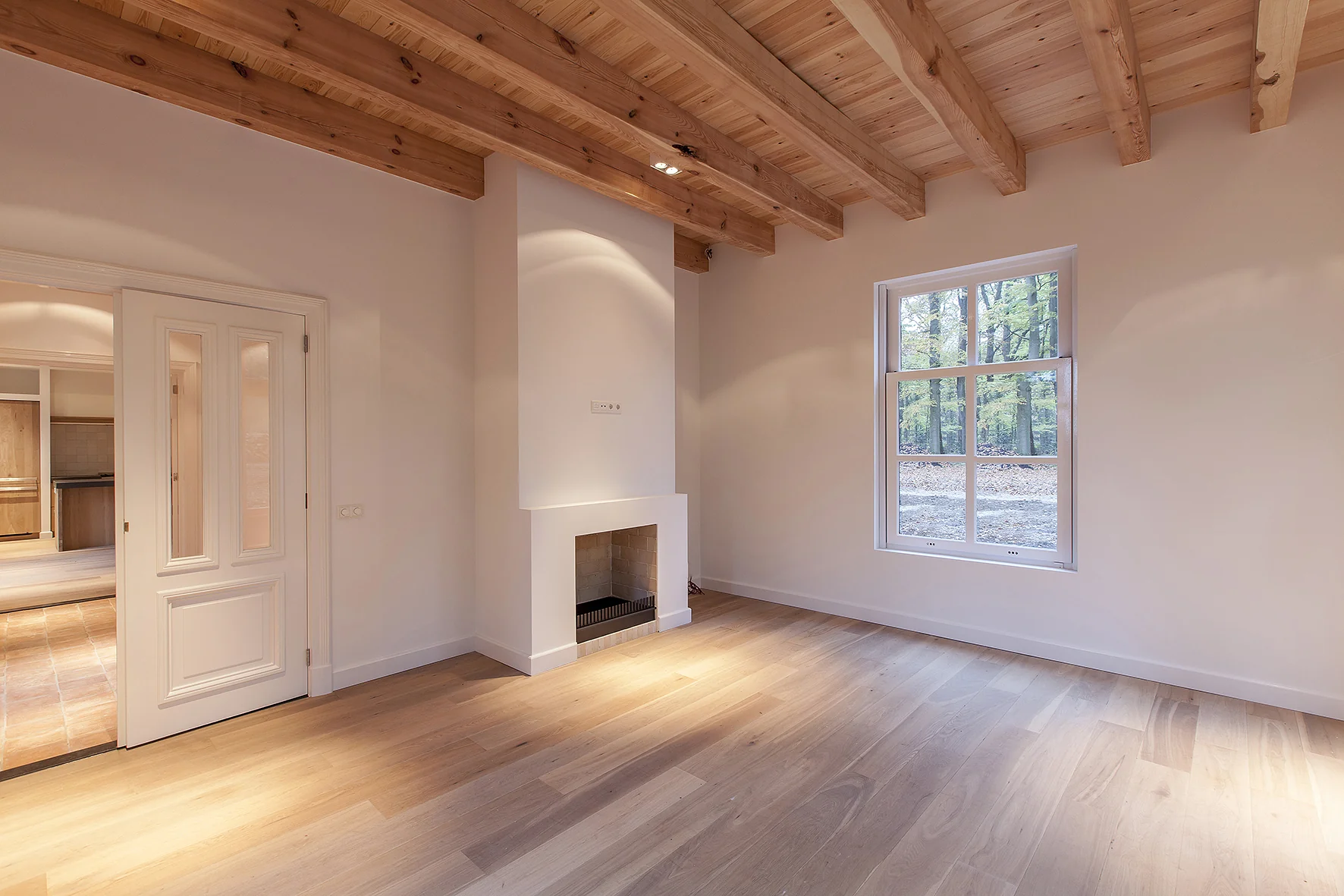Licht en modern interieur met houten vloer, zichtbare balkenplafond, open haard en groot raam met uitzicht op bosrijke omgeving aan de Naarderstraat.