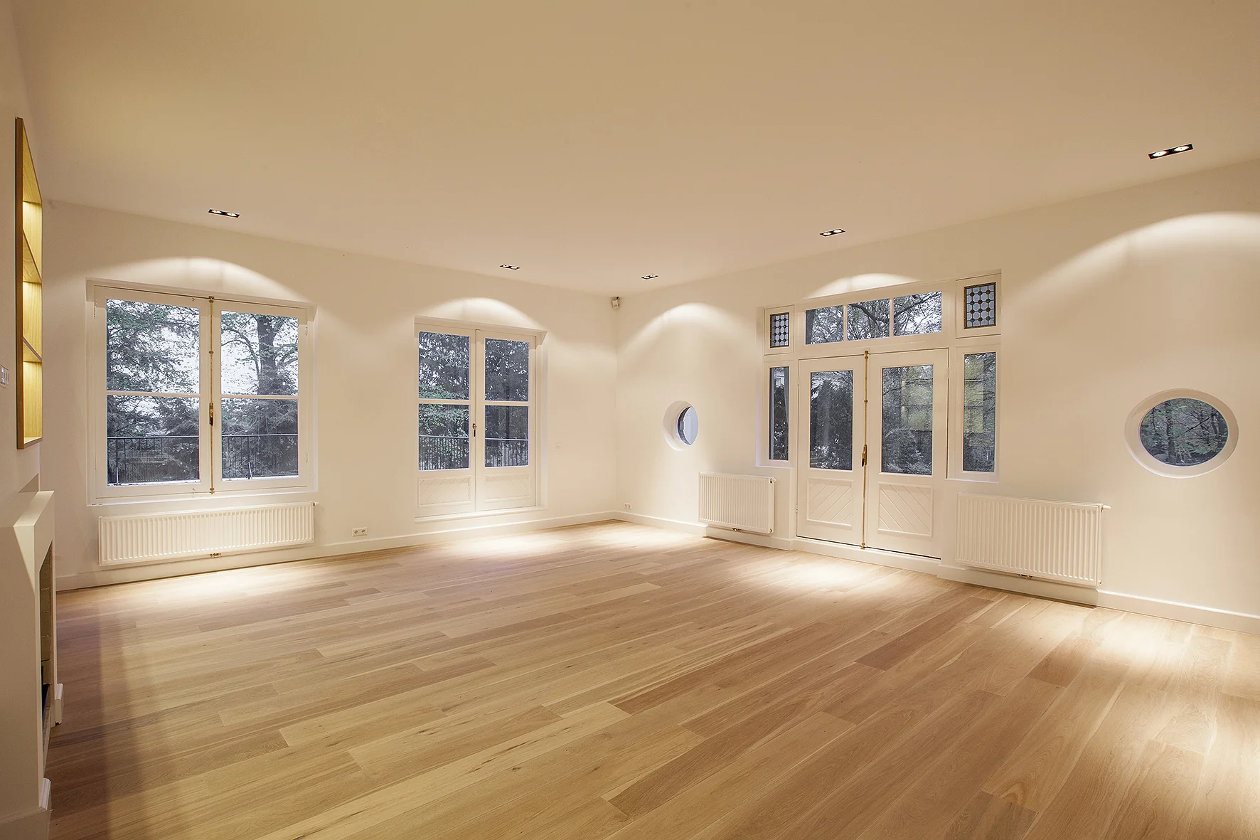 Lichte, lege woonkamer aan de Naarderstraat met houten vloer, grote ramen en openslaande deuren naar buiten.