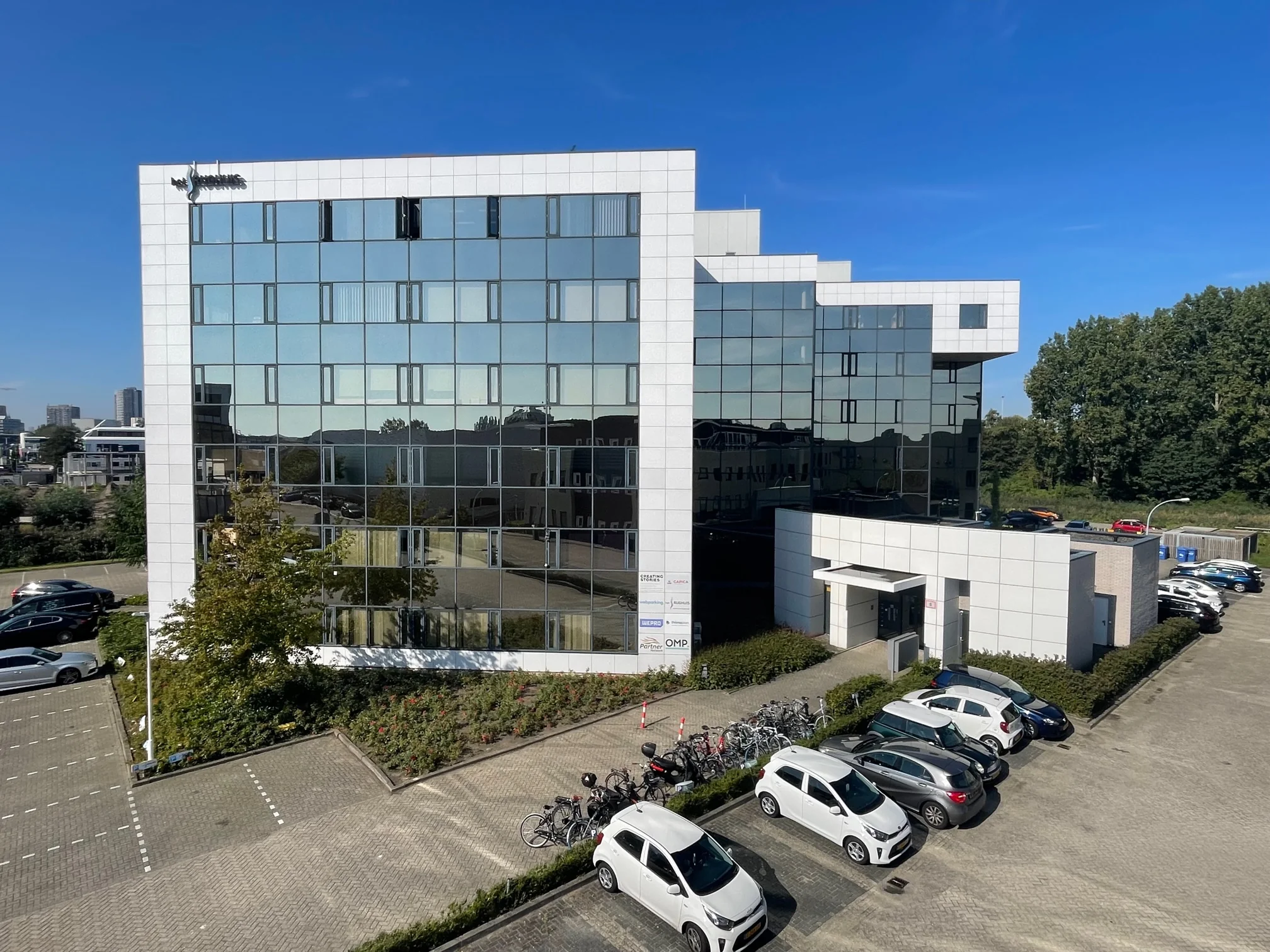 Wit kantoorgebouw aan de Cypresbaan met spiegelramen, fietsenrekken en geparkeerde auto's op de voorgrond.