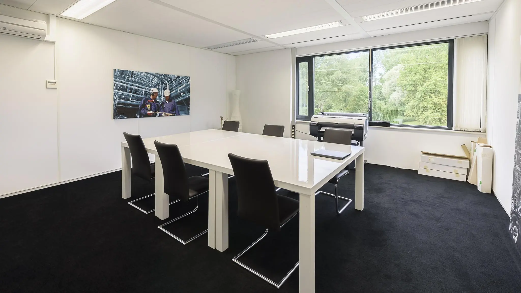 Vergaderruimte met een witte tafel, zes stoelen, groot raam en een foto van industriële werkers aan de muur.