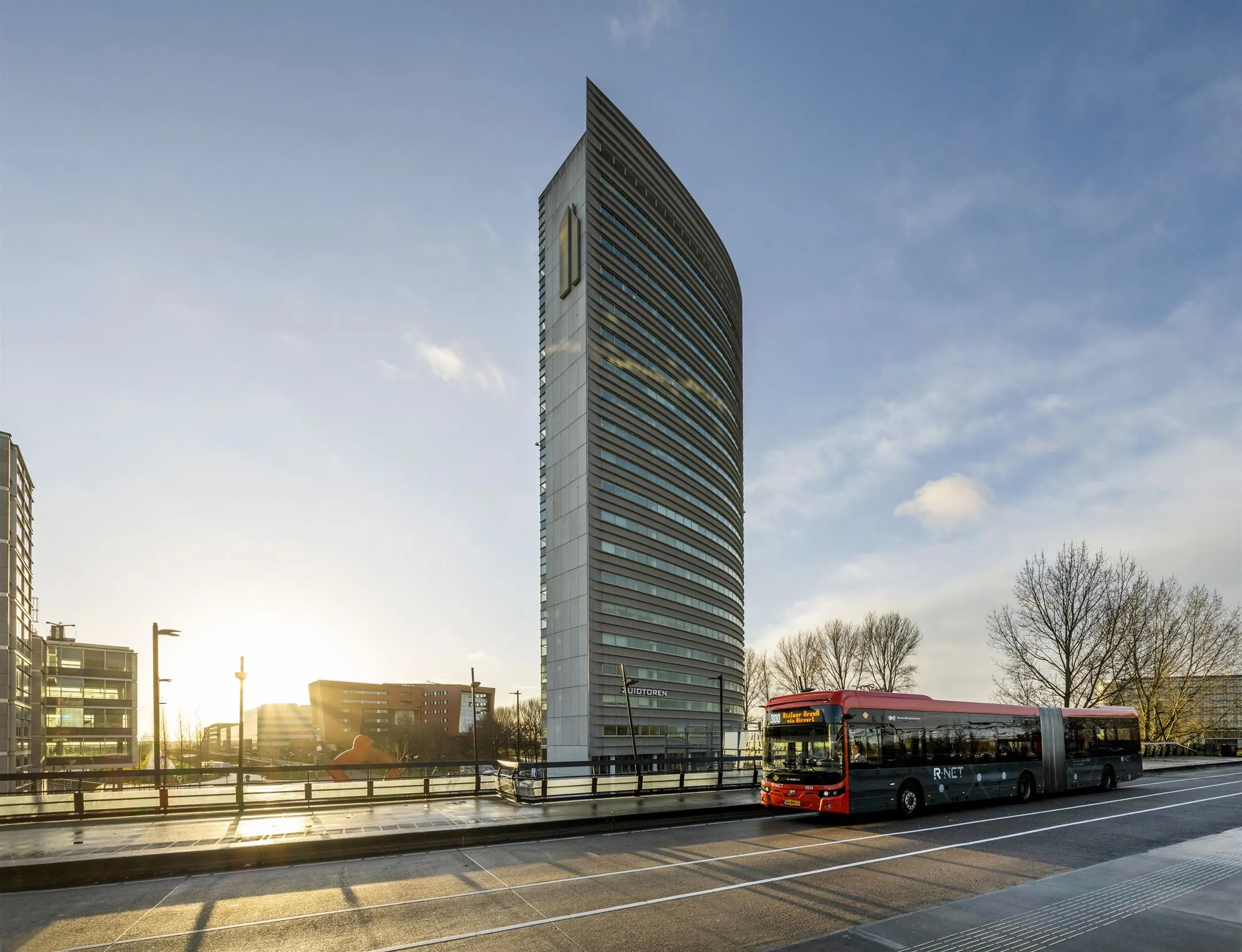 Een rood-grijze R-NET bus staat stil voor het moderne, hoge kantoorgebouw "De Verrekijker" in Zoetermeer bij zonsondergang.