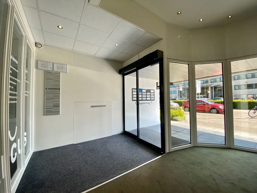 Ingang van een kantoorgebouw aan de Computerweg 11 met glazen draaideur en zicht op geparkeerde auto's buiten.