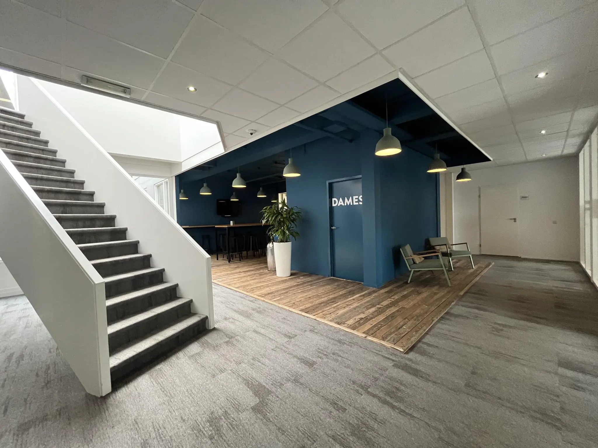Moderne kantoorhal met trap, zithoek en een deur met het opschrift "DAMES" tegen een blauwe wand.