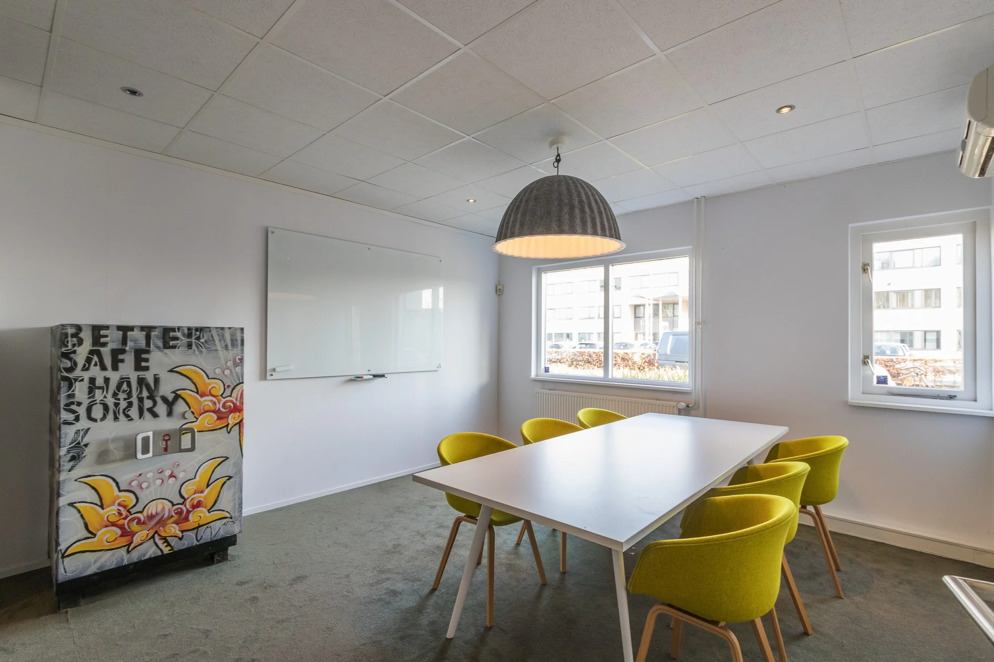 Vergaderruimte met een witte tafel, zes gele stoelen, een whiteboard en een kleurrijke kast met de tekst "Better Safe Than Sorry".