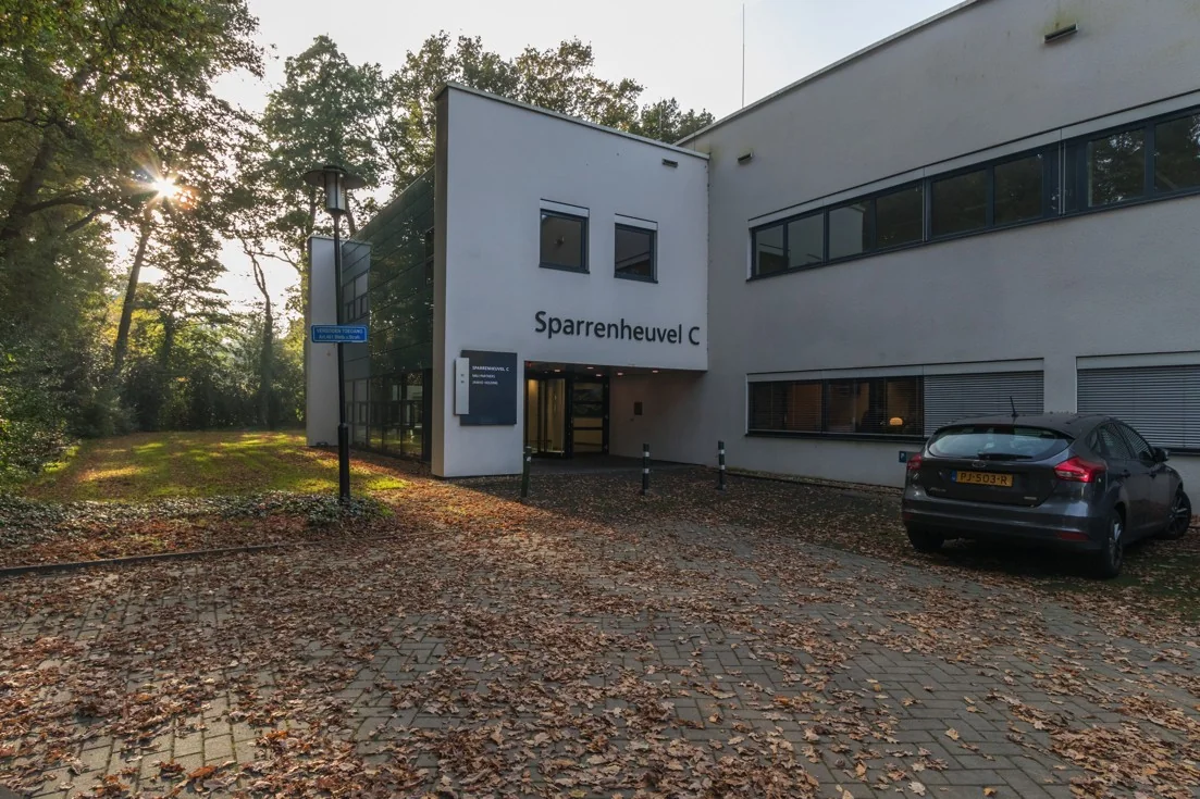 Ingang van het kantoorgebouw Sparrenheuvel C, omgeven door herfstbladeren en geparkeerde auto.