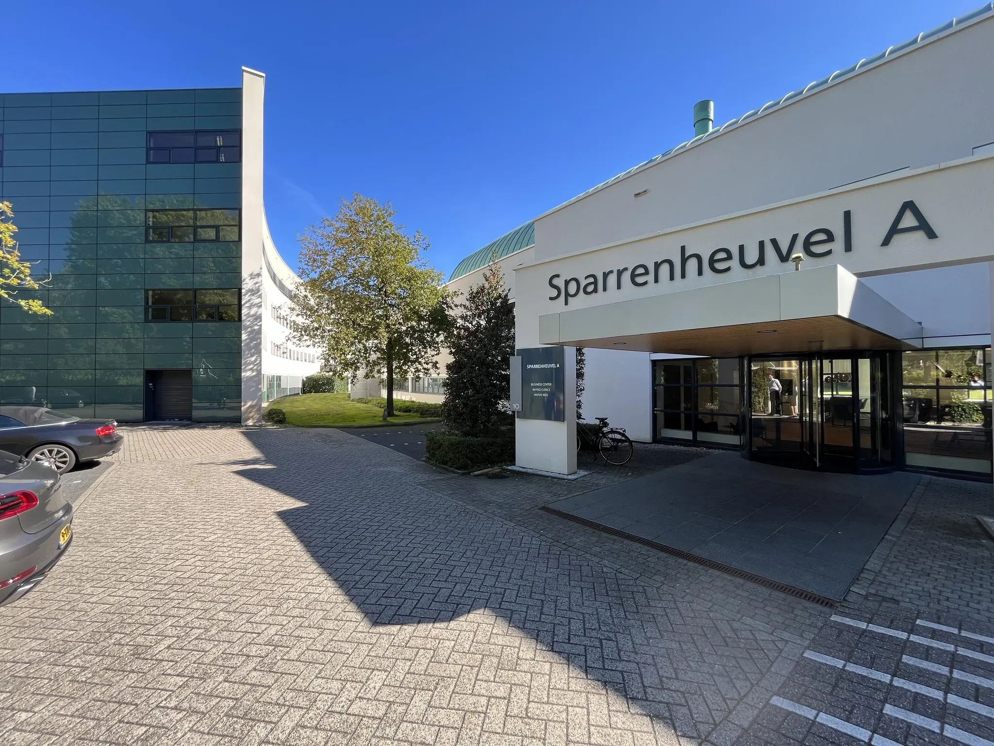 Ingang van het gebouw Sparrenheuvel A met automatische draaideur en omliggende parkeerplaatsen.