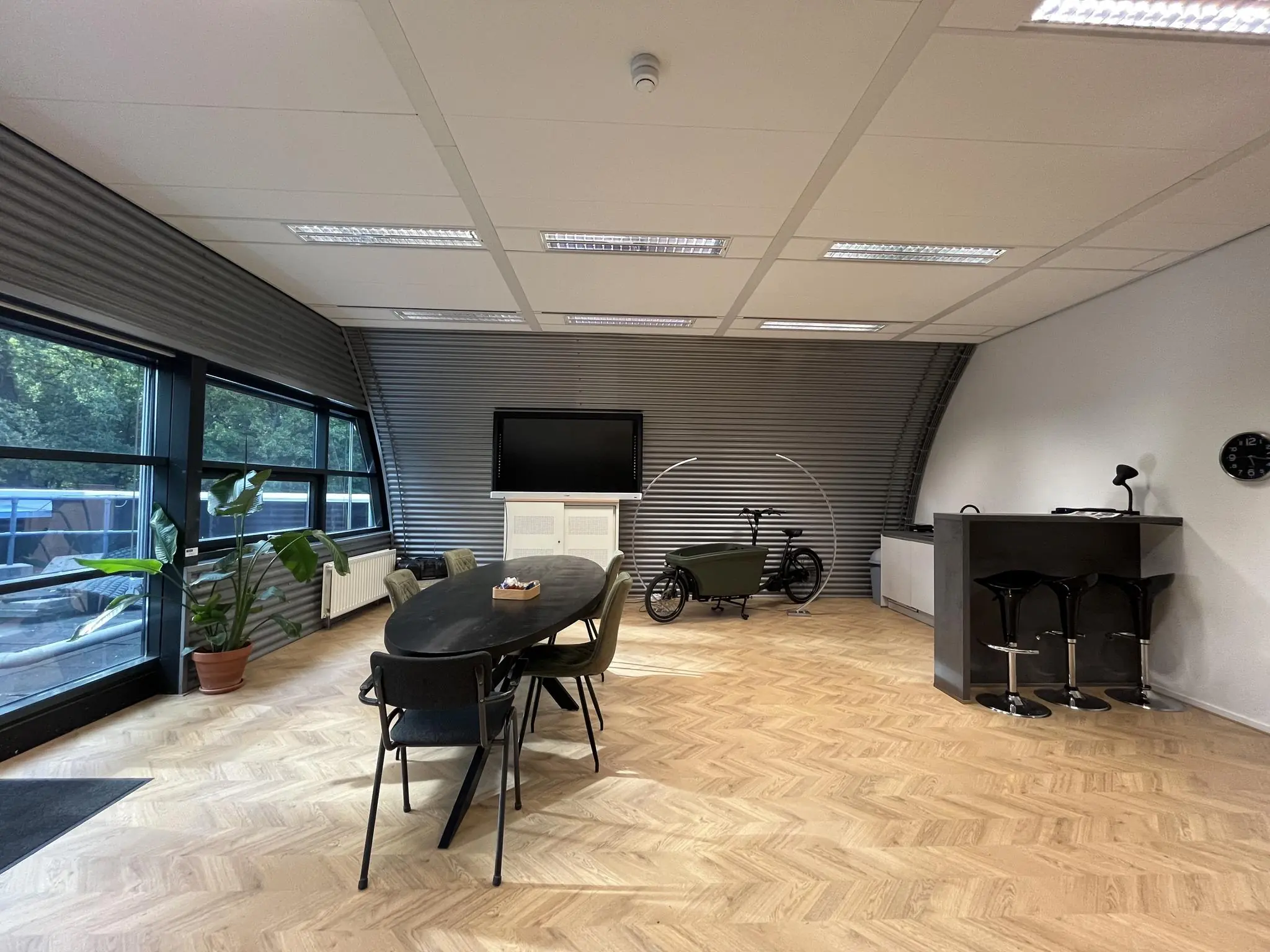 Moderne vergaderruimte op Sparrenheuvel met een ovale tafel, barkrukken, een bakfiets en grote ramen met uitzicht op groen.