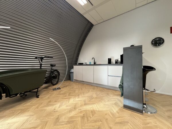 Moderne pantryruimte in kantoor Sparrenheuvel met houten visgraatvloer, designkeuken en bakfiets tegen een metalen wand.