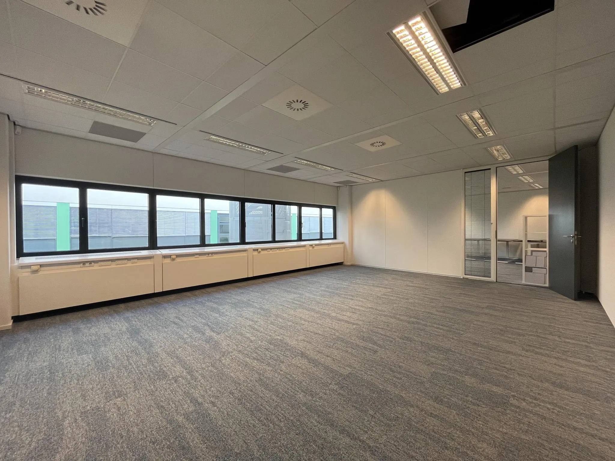 Licht en ruim leeg kantoor op de Sparrenheuvel met grote ramen en grijze vloerbedekking.