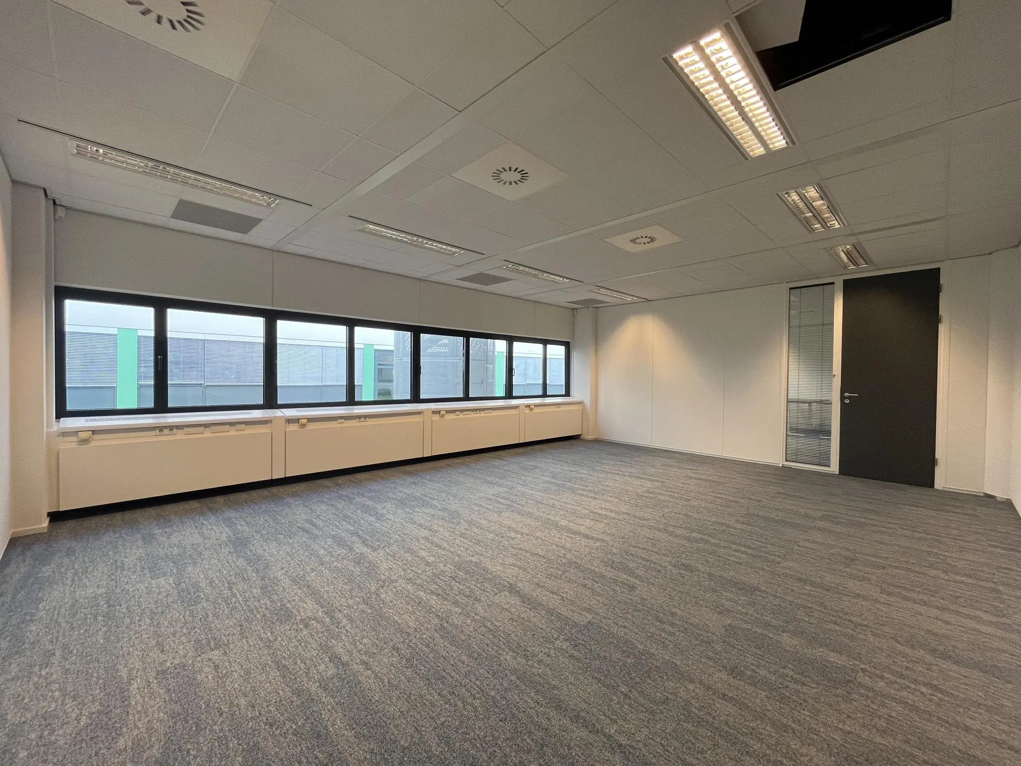Lege kantoorruimte met grijs tapijt, witte muren, grote ramen en systeemplafond in gebouw Sparrenheuvel.