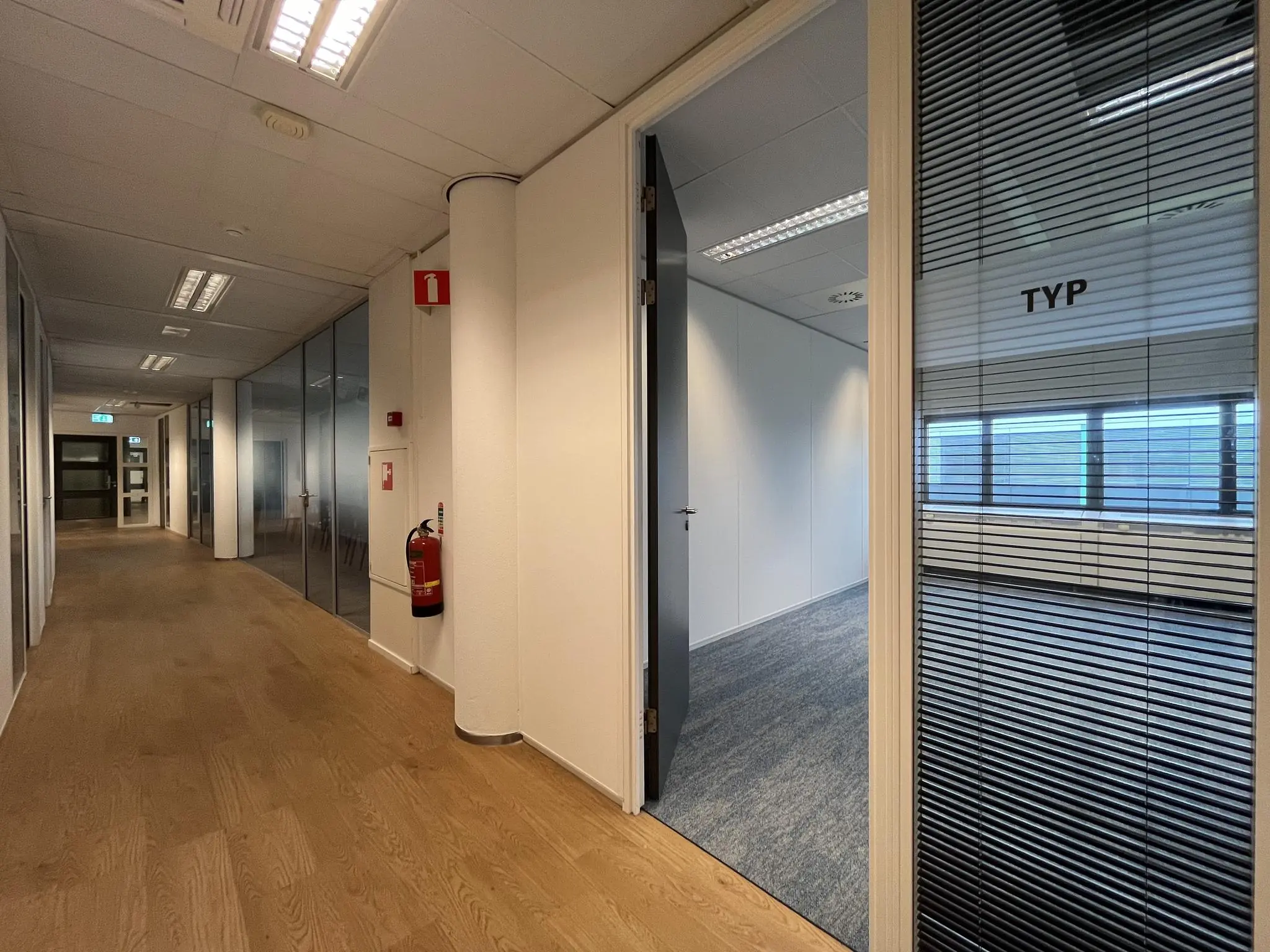 Moderne kantoorgang met houten vloer, glazen wanden en een open deur naar vergaderruimte 'TYP' in het gebouw Sparrenheuvel.