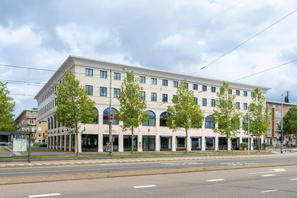 Modern hoekgebouw aan het Stadionplein in Amsterdam met bomenrij en tramhalte op de voorgrond.
