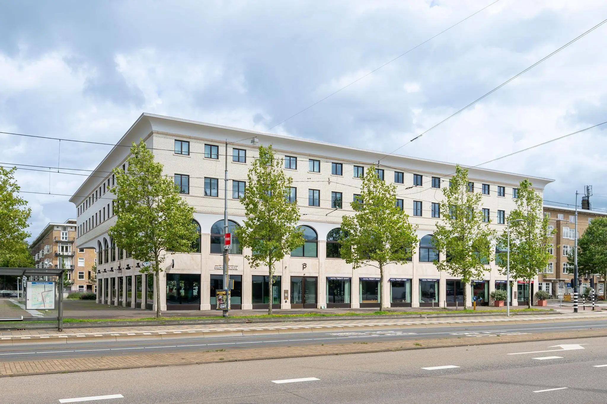 Modern hoekgebouw aan het Stadionplein in Amsterdam met bomenrij en tramhalte op de voorgrond.