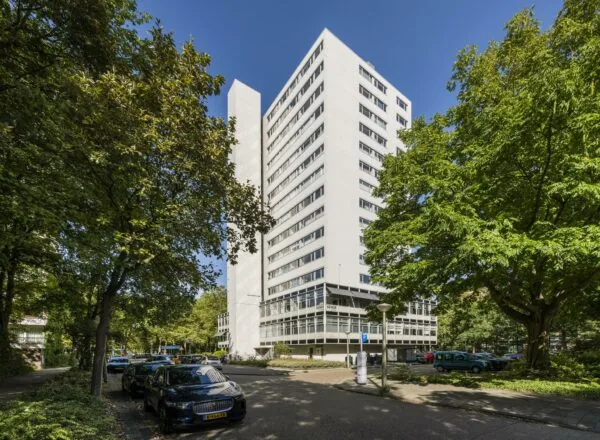 Wit woongebouw Backershagen in Den Haag omringd door bomen en geparkeerde auto’s op een zonnige dag.