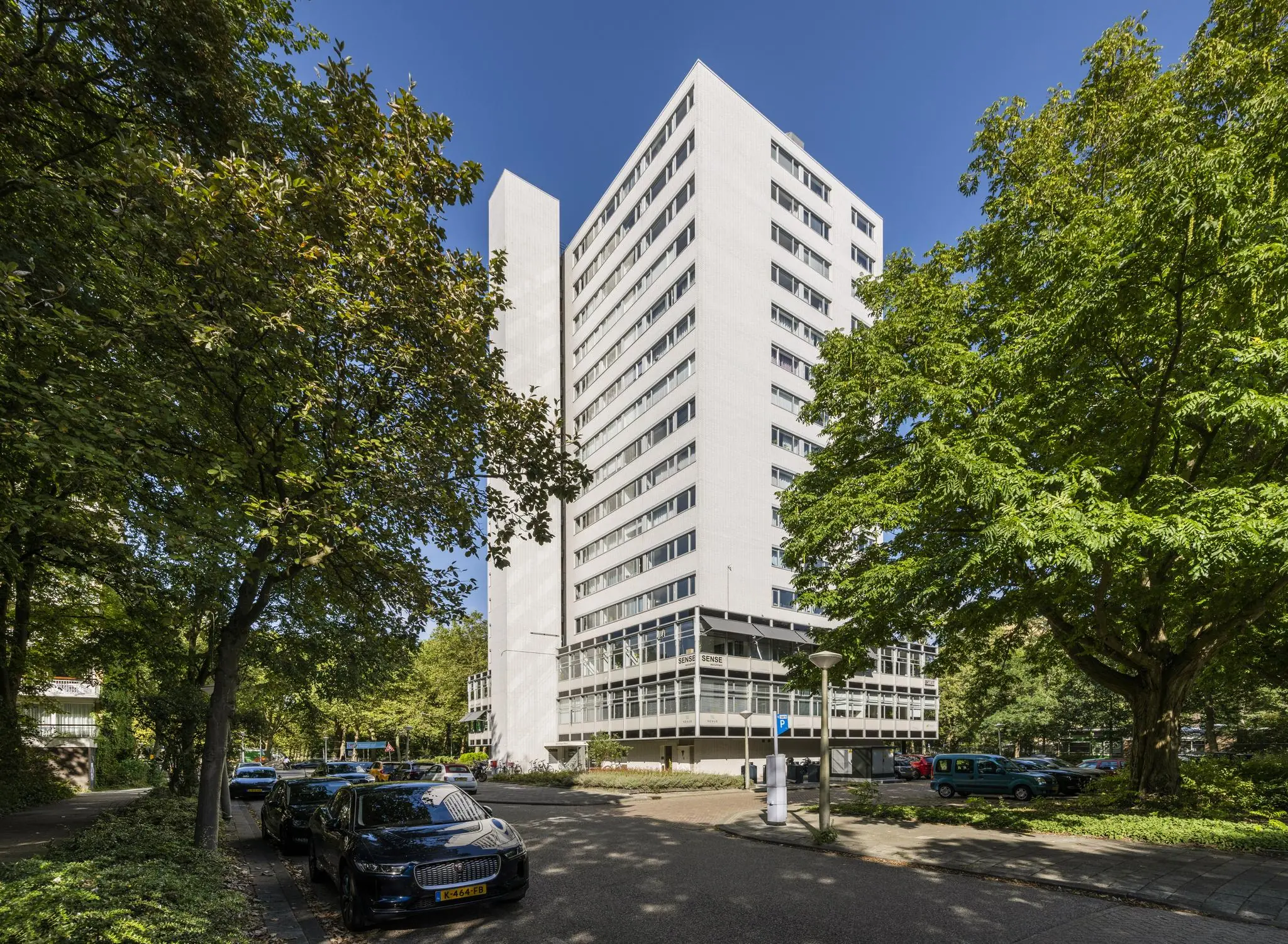Wit woongebouw Backershagen in Den Haag omringd door bomen en geparkeerde auto’s op een zonnige dag.