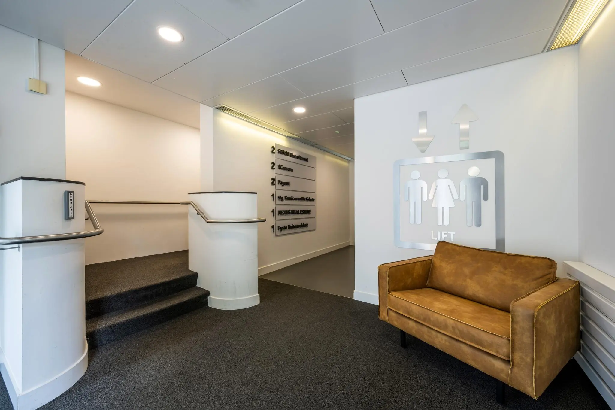 Wachtruimte met een bruine bank, liftpictogram op de muur en bewegwijzering naar bedrijven in verzamelpand Backershagen.