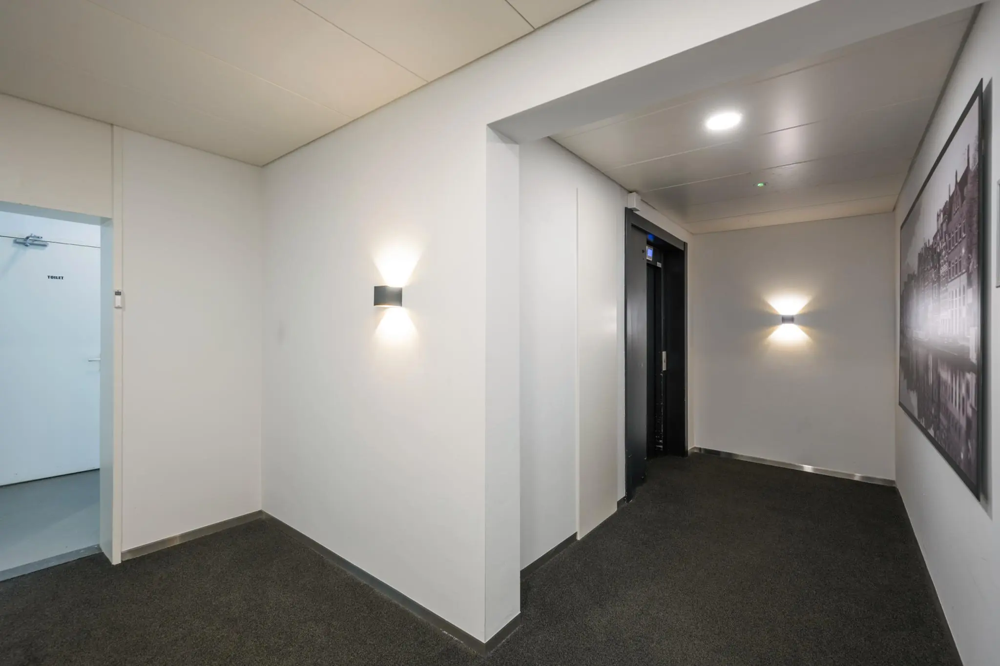 Hal met een liftdeur, een open toiletruimte en wandverlichting in het gebouw Backershagen.