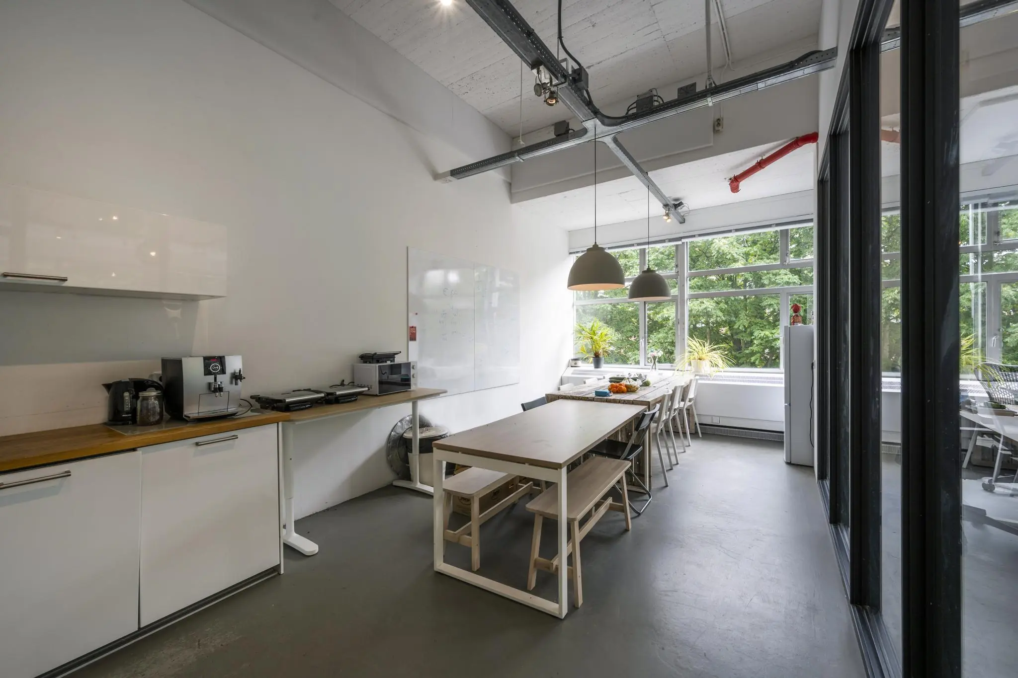 Moderne, lichte kantoorkeuken met lange houten eettafel, koffieapparaat en uitzicht op groen.