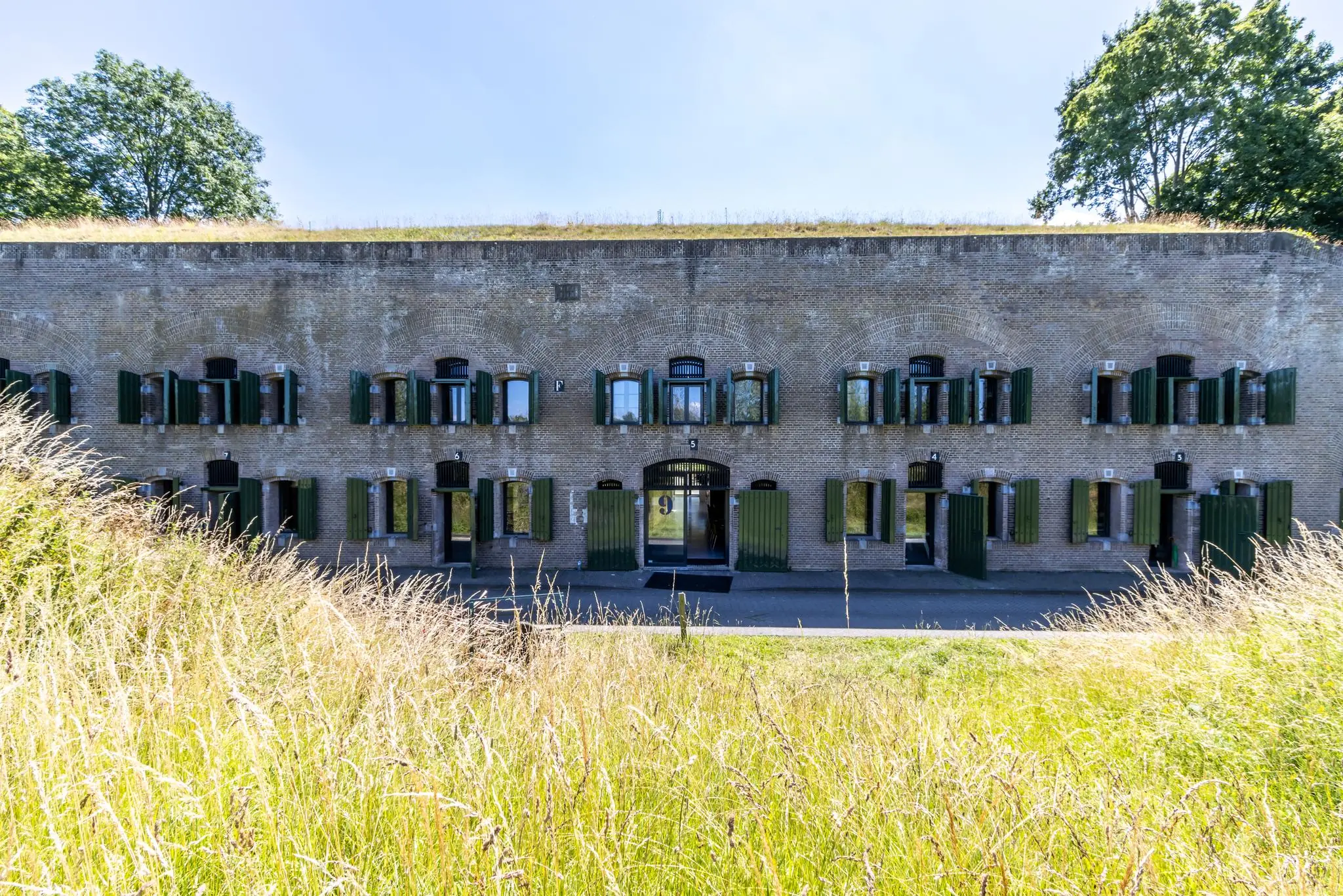 Vooraanzicht van een historisch fortgebouw aan de Fortweg met openstaande groene luiken en een grasveld op de voorgrond.