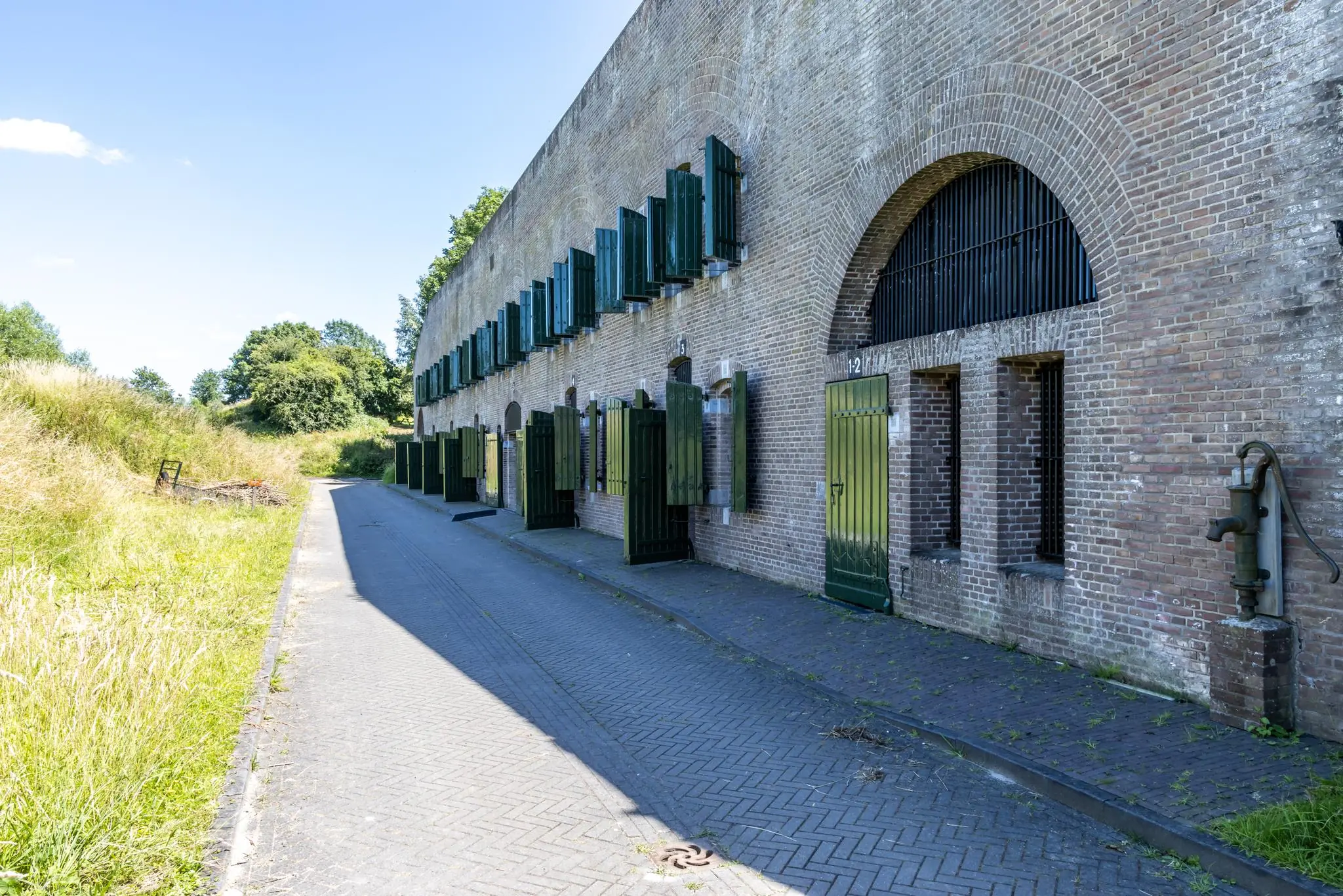 Bakstenen fortgebouw aan de Fortweg met groene deuren en luiken langs een smal geplaveid pad.