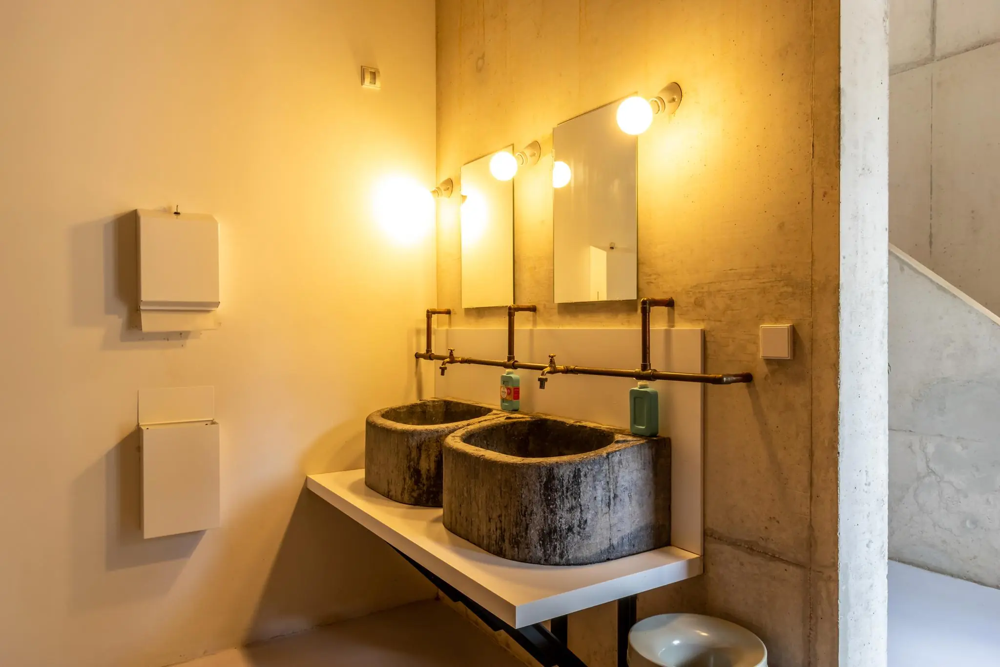 Industriële badkamer met twee betonnen wasbakken, spiegels en zichtbare koperen leidingen.