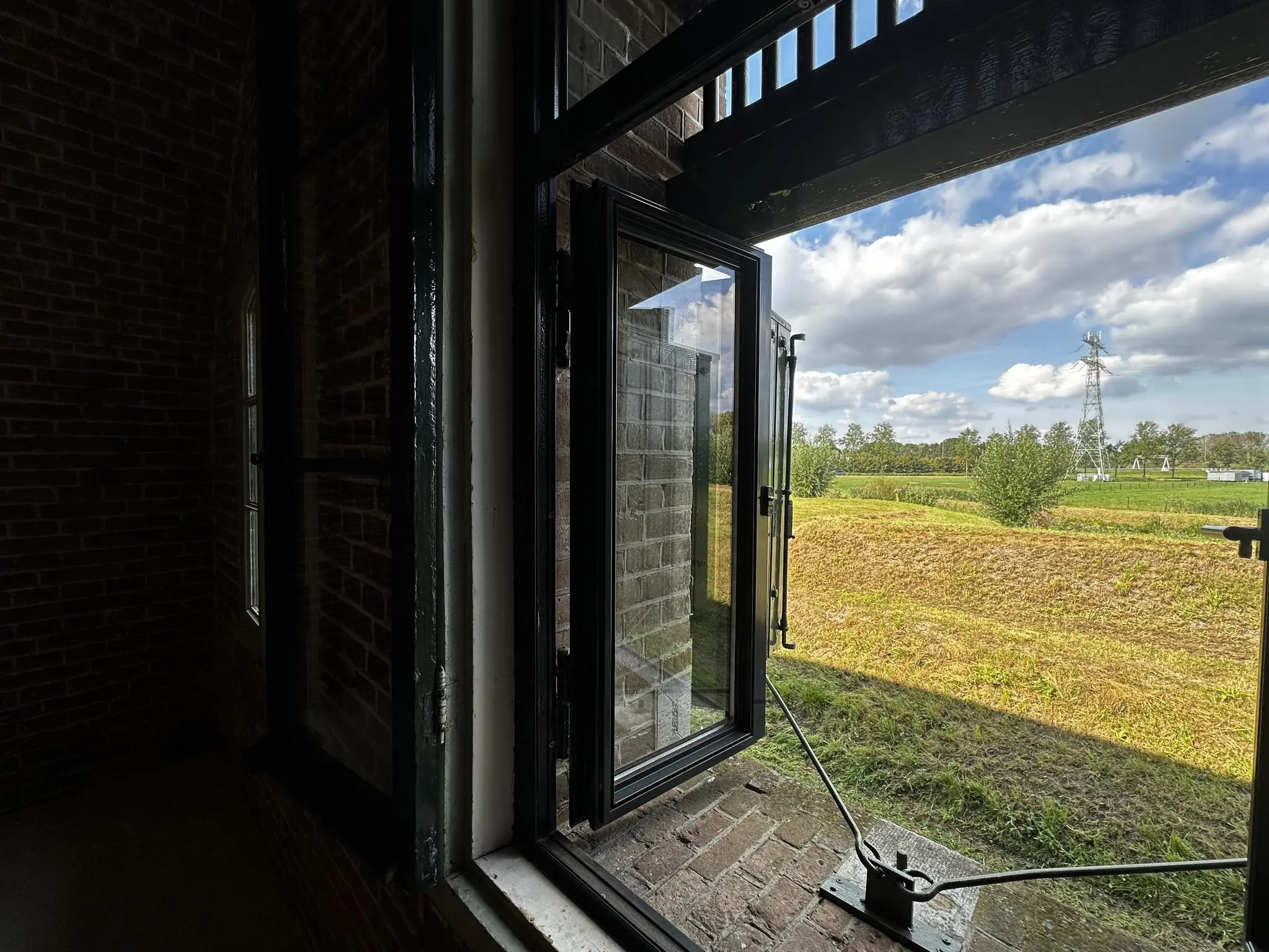 Uitzicht vanuit een open raam van een oud fortgebouw aan de Fortweg op een groen landschap met bomen en een hoogspanningsmast.