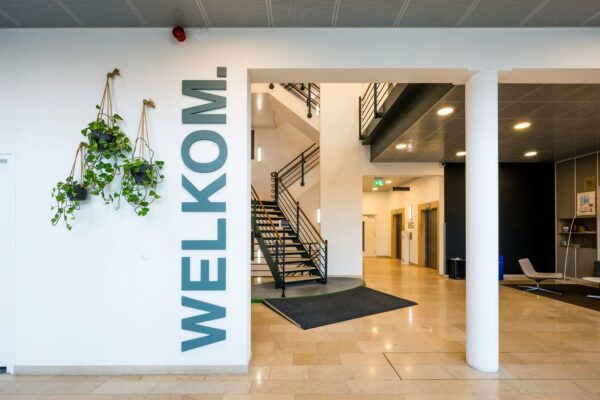 Ingang van een modern kantoorpand aan de Watermanweg met een welkom-tekst op de muur, hangplanten en een centrale trap.