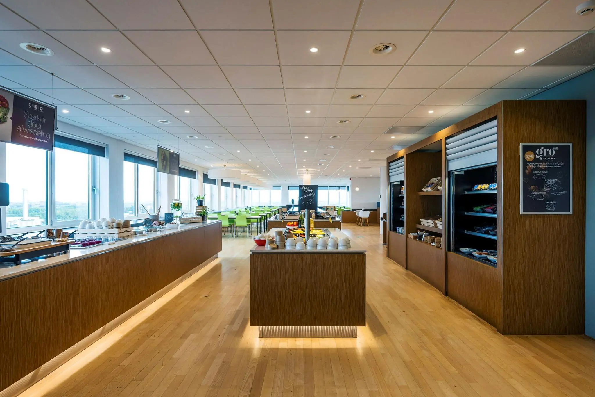 Moderne bedrijfsrestaurant aan de Watermanweg met houten vloer, zelfbedieningsbuffet en groene stoelen bij grote ramen.