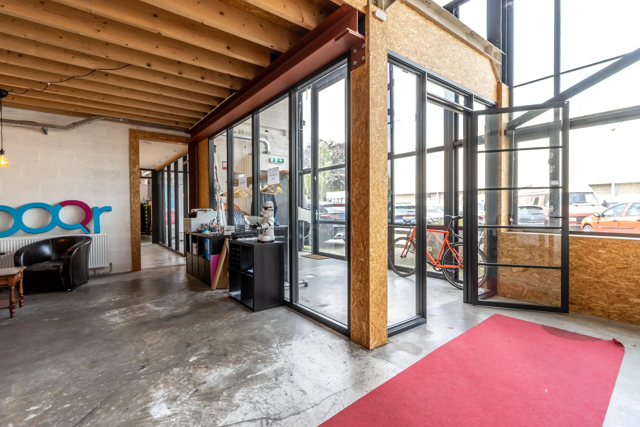 Ingang van een modern kantoor aan de Eendrachtlaan met glazen deuren, een rode loper en een oranje fiets buiten.