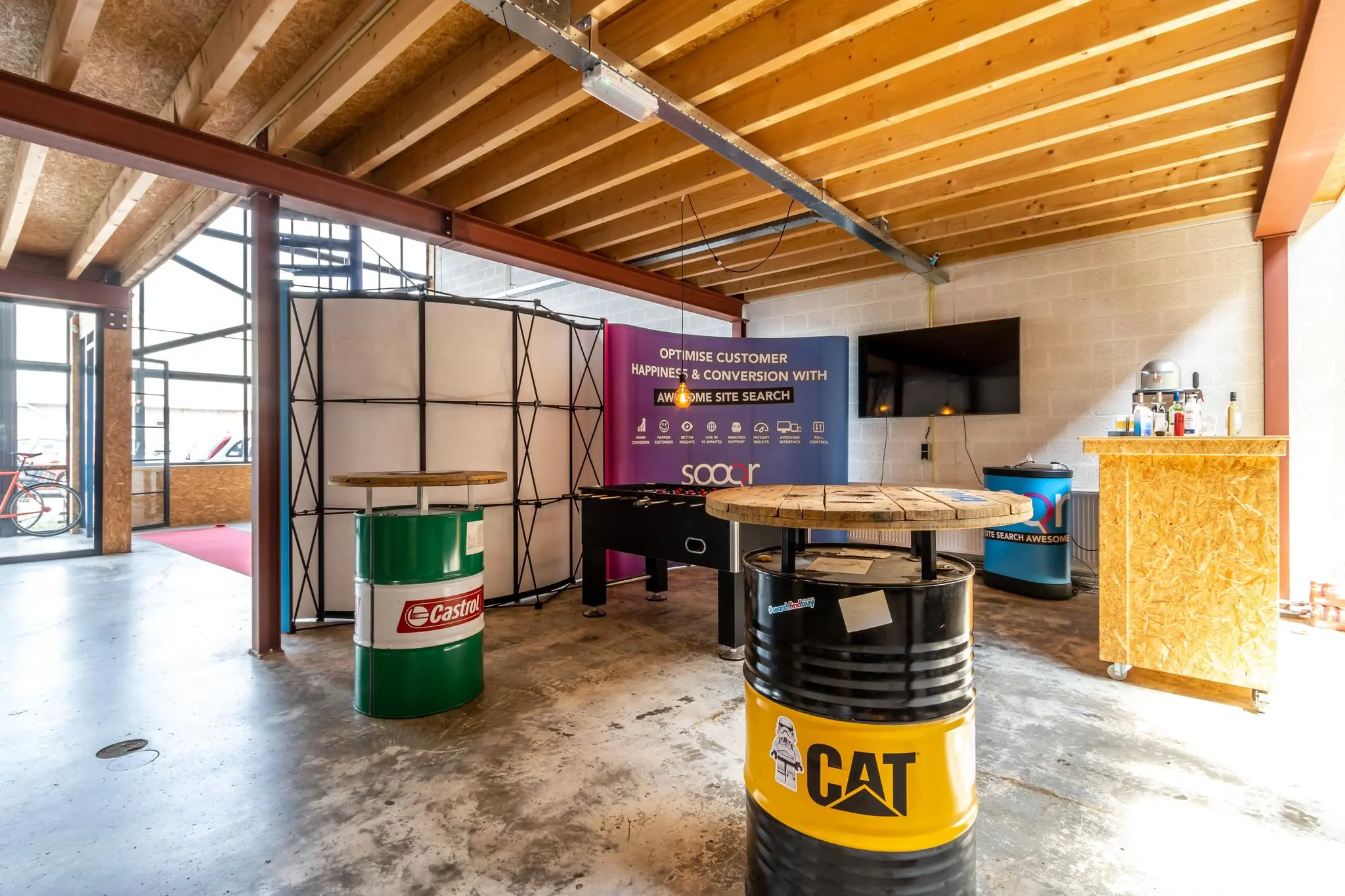 Industrieel ingerichte recreatieruimte met olievat-tafels, een tafelvoetbalspel en een banner van het bedrijf Sooqr in een pand aan de Eendrachtlaan.