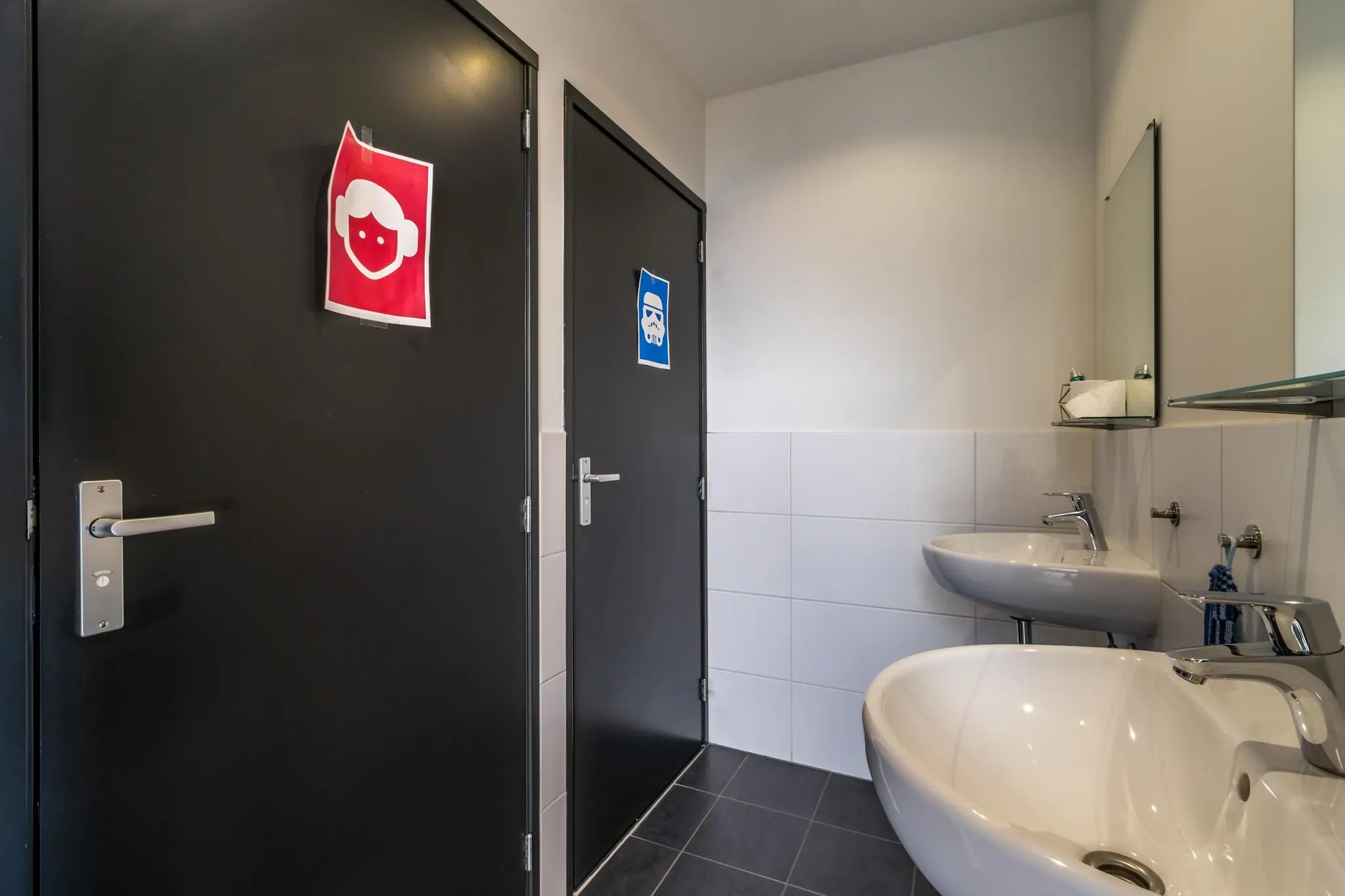 Twee toiletten met zwarte deuren en speelse pictogrammen, naast een wand met twee wastafels en een spiegel, in het gebouw aan de Eendrachtlaan.