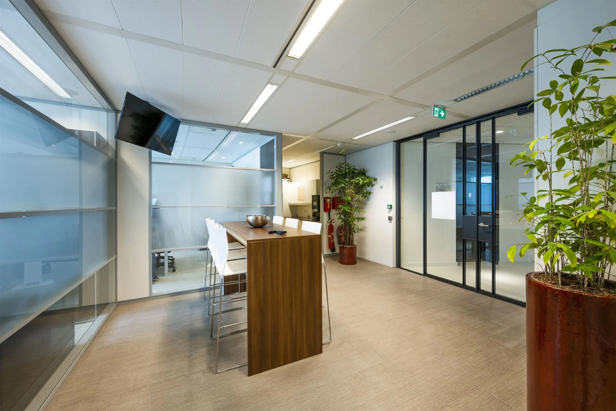 Moderne kantoorlounge met hoge houten tafel, witte barkrukken, glazen wanden en groene kamerplanten.