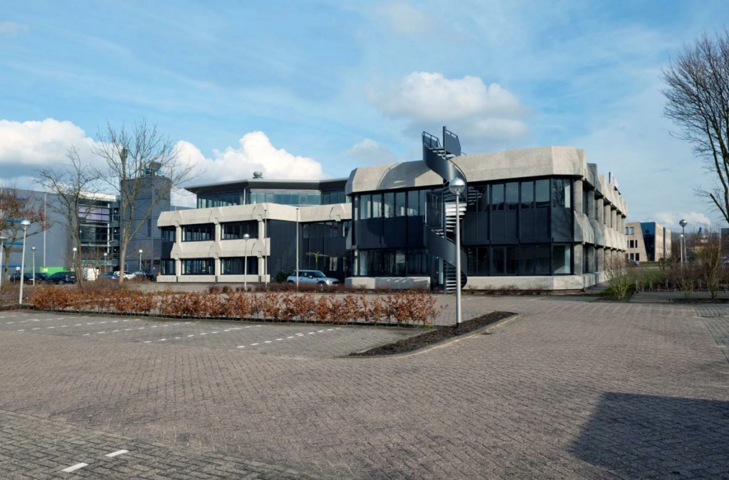 Kantoorgebouw aan de Wattbaan met opvallende betonnen gevel en buitentrap, gezien vanaf een lege parkeerplaats.