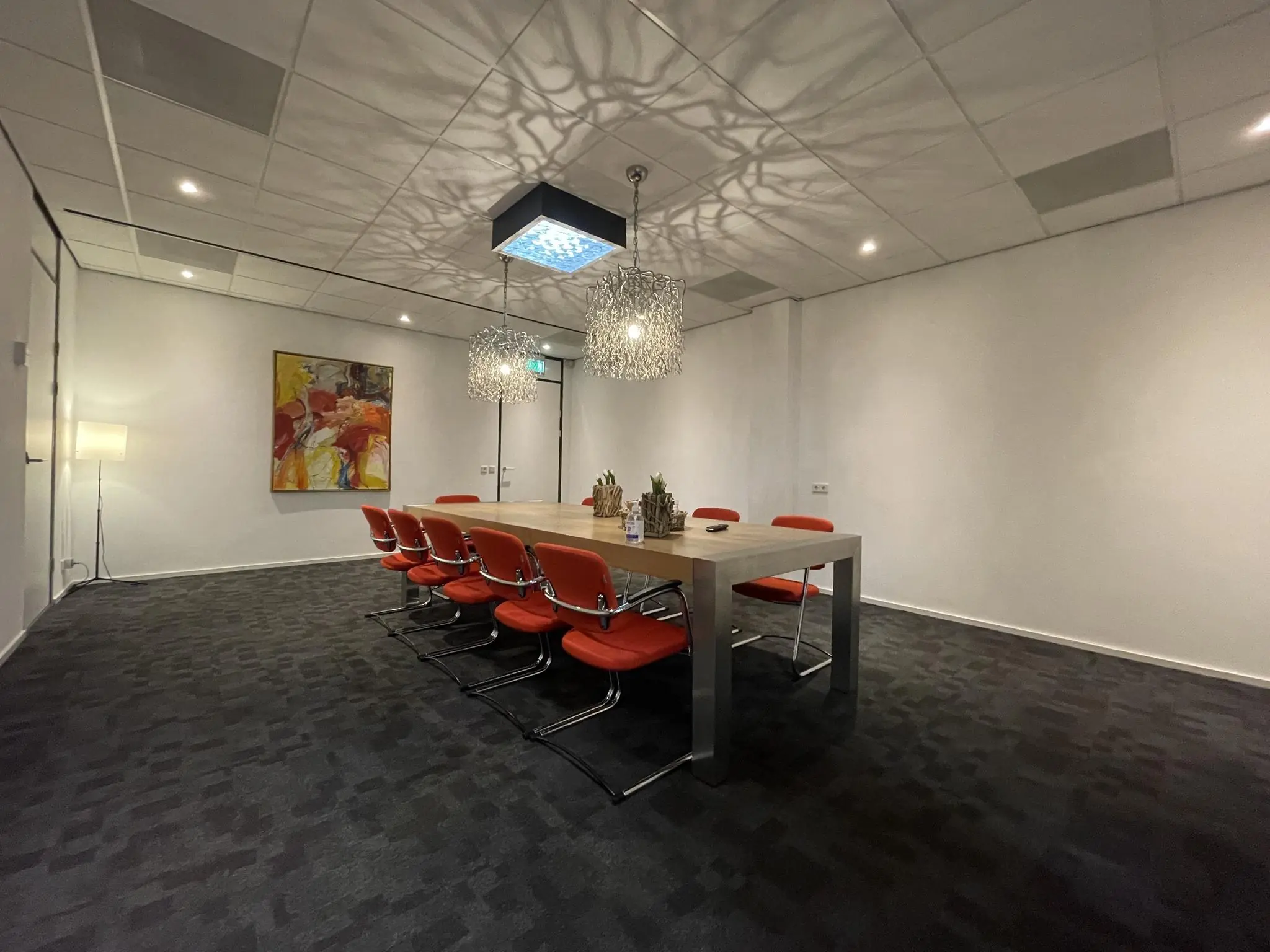 Vergaderruimte met een lange houten tafel, rode stoelen, moderne verlichting en een abstract schilderij aan de muur.