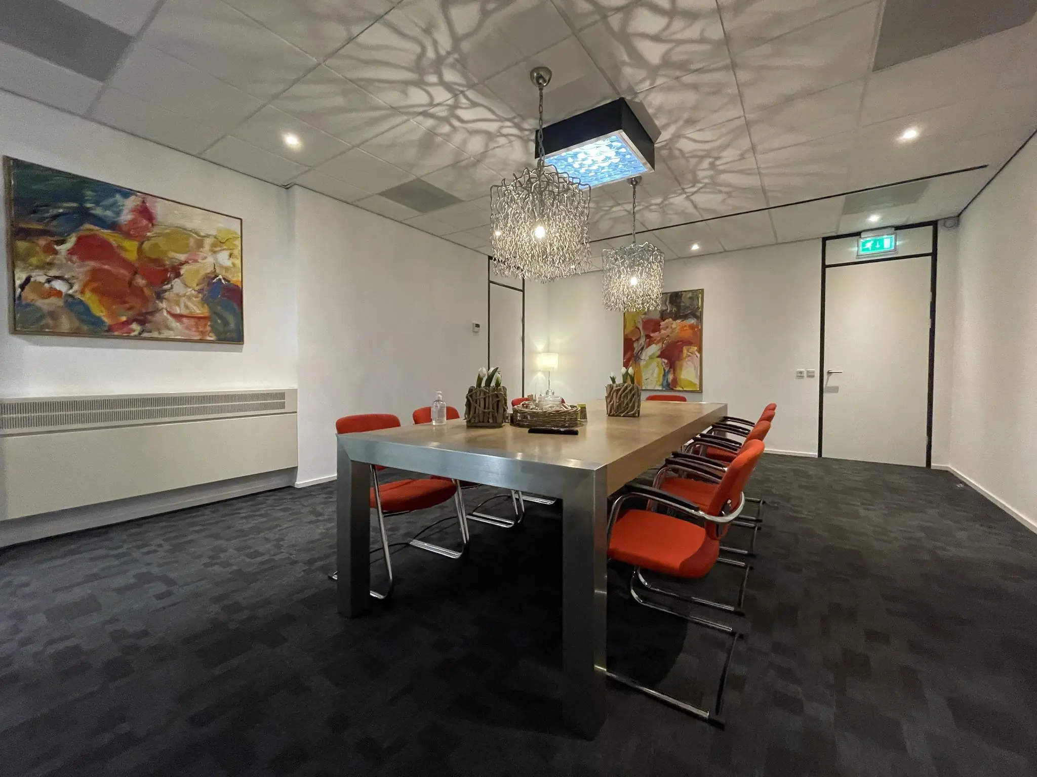 Vergaderruimte met een grote houten tafel, zes oranje stoelen, moderne verlichting en kleurrijke kunst aan de muren.