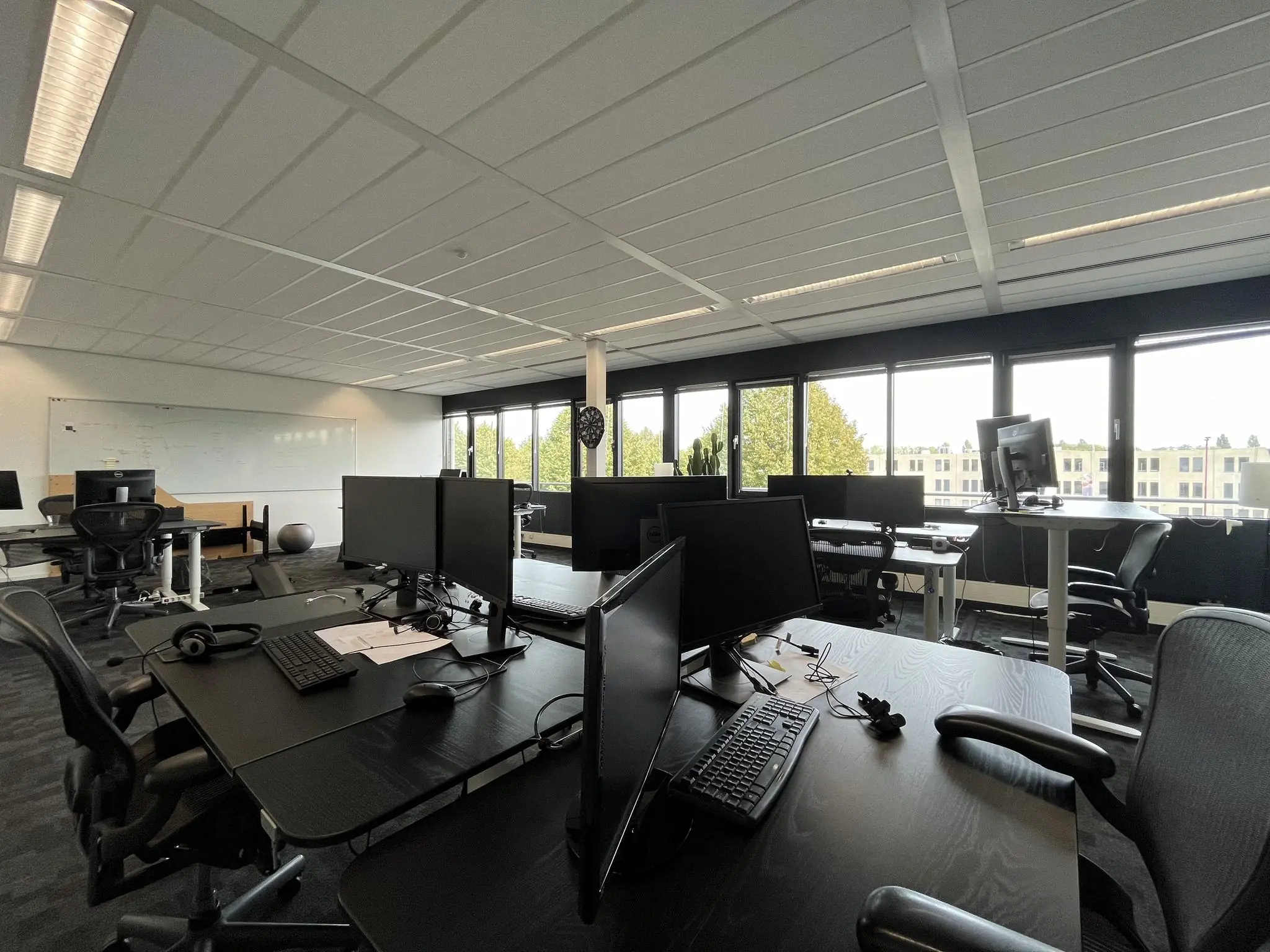 Moderne open kantoorruimte aan de Wattbaan met bureaus, computerschermen en grote ramen met uitzicht op bomen en gebouwen.