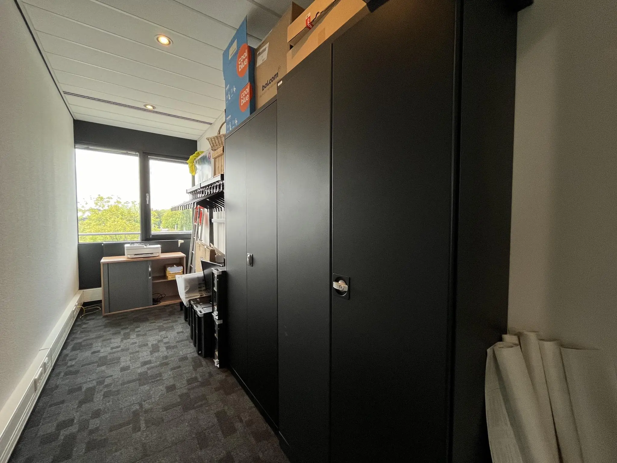 Smalle kantoorruimte met zwarte opbergkasten, een printertafel bij het raam en diverse kantoormaterialen.
