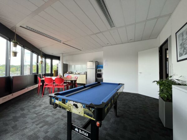 Moderne kantoorruimte met een blauwe pooltafel, zithoek met rode stoelen en een kitchenette op de achtergrond.