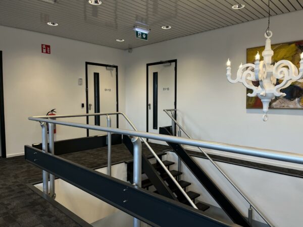 Trapopgang met metalen leuningen in een kantoorpand aan de Wattbaan, met zicht op deuren, kunstwerk en een witte decoratieve lamp.