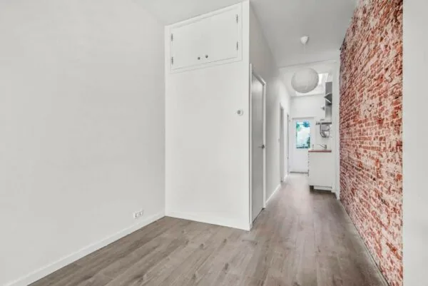Lichte gang in een woning aan de Westerstraat met houten vloer, witte muren en een opvallende bakstenen accentmuur.