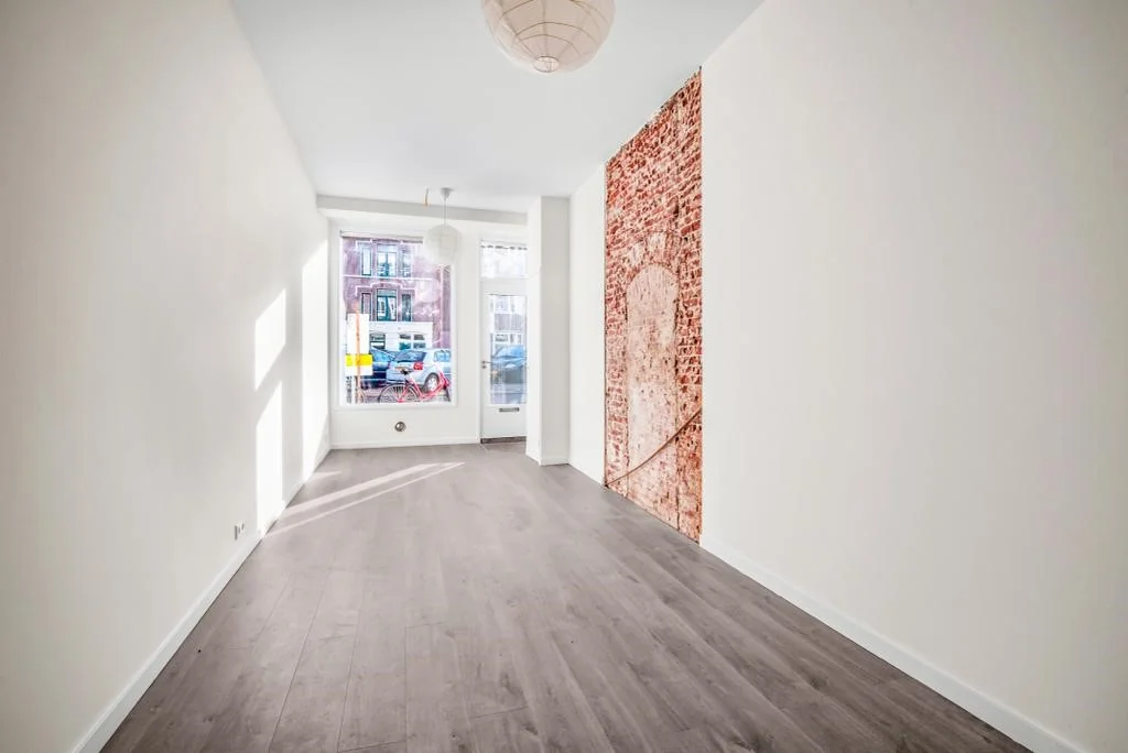 Lichte, smalle kamer met houten vloer, witgestucte muren en een decoratieve bakstenen muurfoto aan de rechterzijde, gelegen aan de Westerstraat.