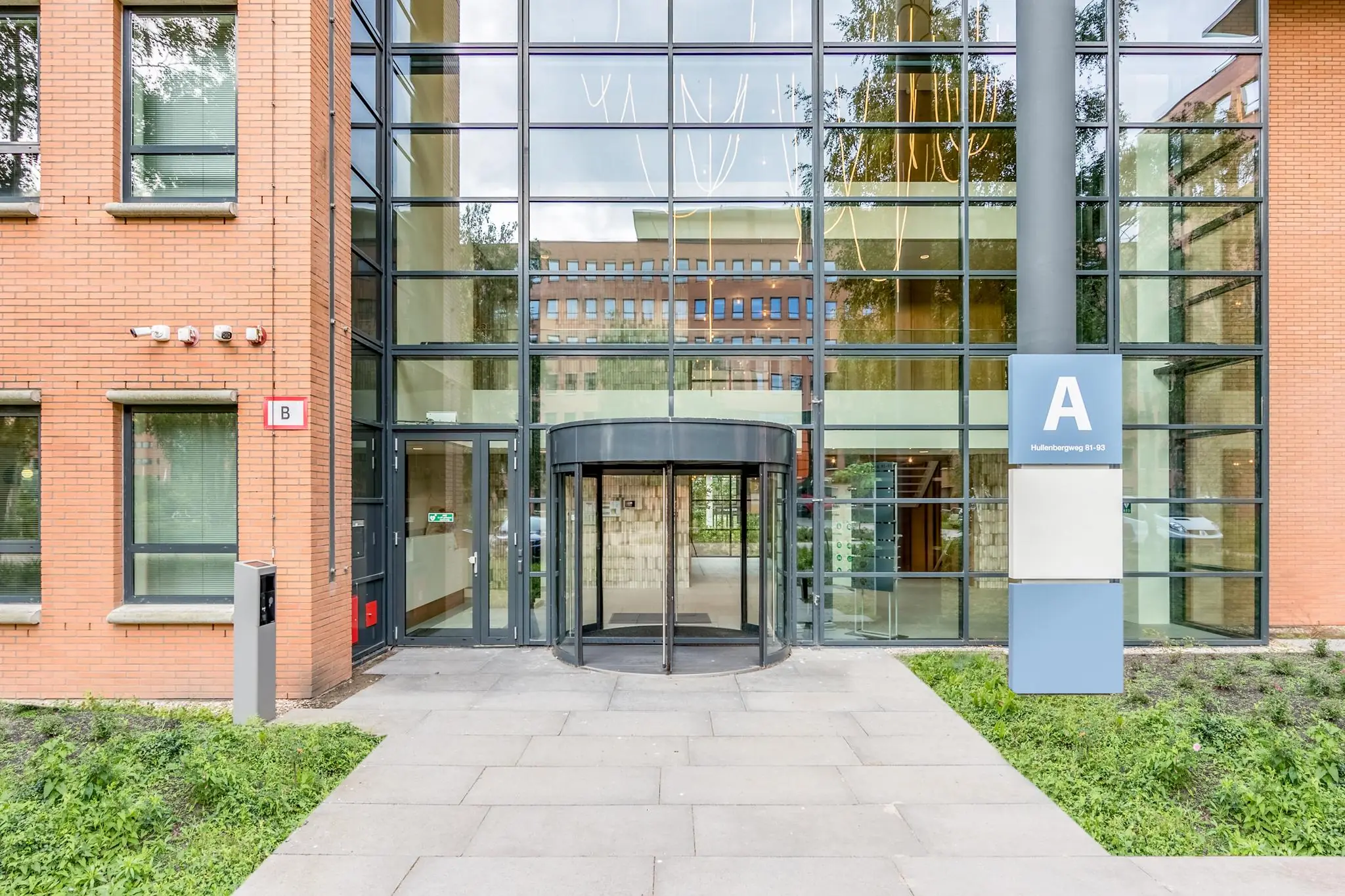 Ingang van een kantoorgebouw aan de Hullenbergweg met een grote glazen gevel, een draaideur en aanduidingen voor gebouwen A en B.