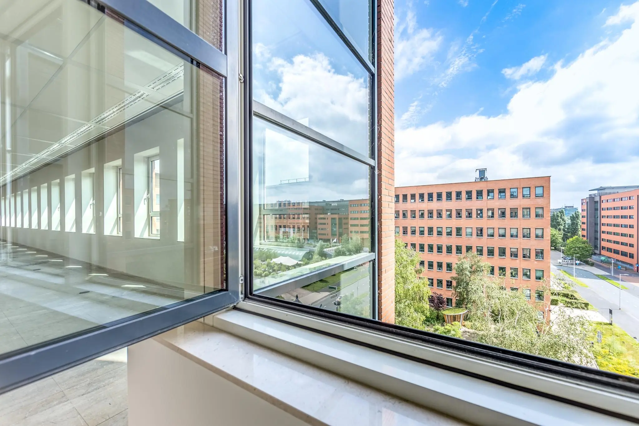 Uitzicht vanuit een open raam op de Hullenbergweg met omliggende moderne kantoorgebouwen en een heldere blauwe lucht.