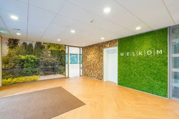 Moderne entree met houten vloer, groene mossige wand met "WELKOM" en fotowand van de toegangspoort tot Amaliapark.