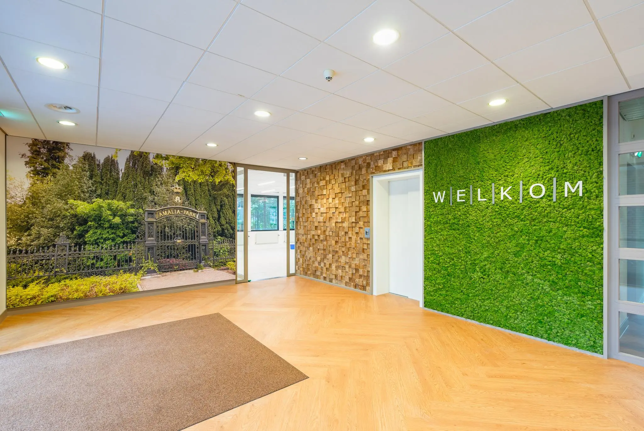 Moderne entree met houten vloer, groene mossige wand met "WELKOM" en fotowand van de toegangspoort tot Amaliapark.