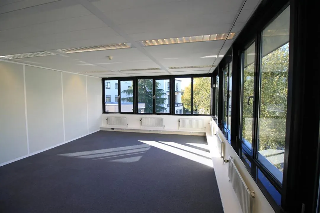 Lichte, lege kantoorruimte aan de Amaliastraat met grote ramen en uitzicht op groene bomen.