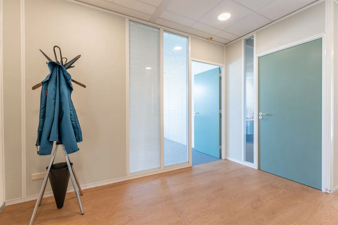 Hal met houten vloer, kapstok met blauwe jas en deuren naar aangrenzende kantoorruimtes aan de Amalialaan.