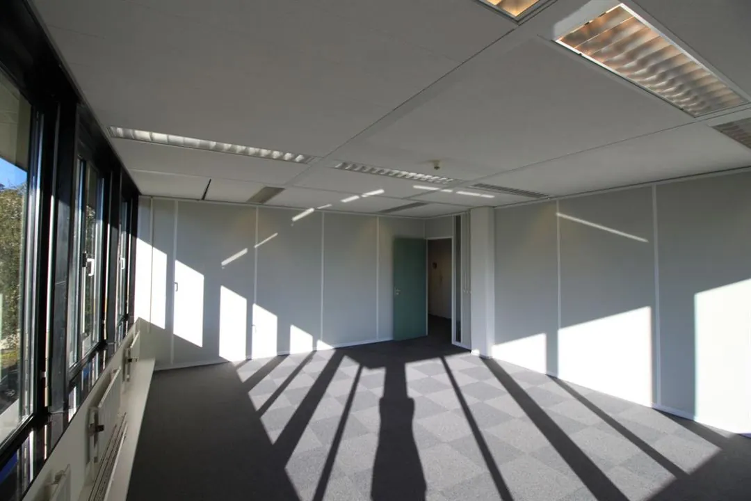 Licht en leeg kantoor aan de Amaliastraat met witte wanden, grijs tapijt en zonlicht dat door de ramen valt.