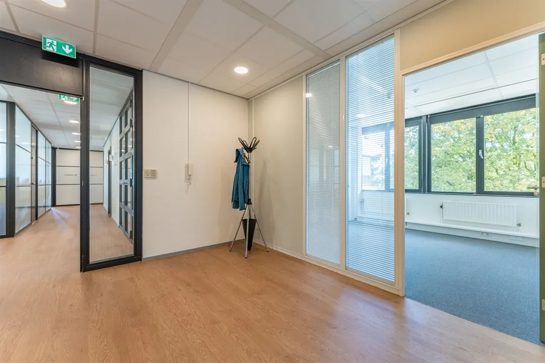 Entree van een modern kantoor aan de Amalialaan met glazen wanden, houten vloer en zicht op een aangrenzend kantoor met grote ramen.